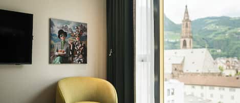 Habitación de hotel con cama, una silla, una mesita, un cuadro y vista a un paisaje urbano con una torre de iglesia.