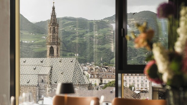 Une table à manger avec des chaises et un vase de fleurs, donnant sur un paysage urbain où l’on aperçoit une église imposante et des collines.
