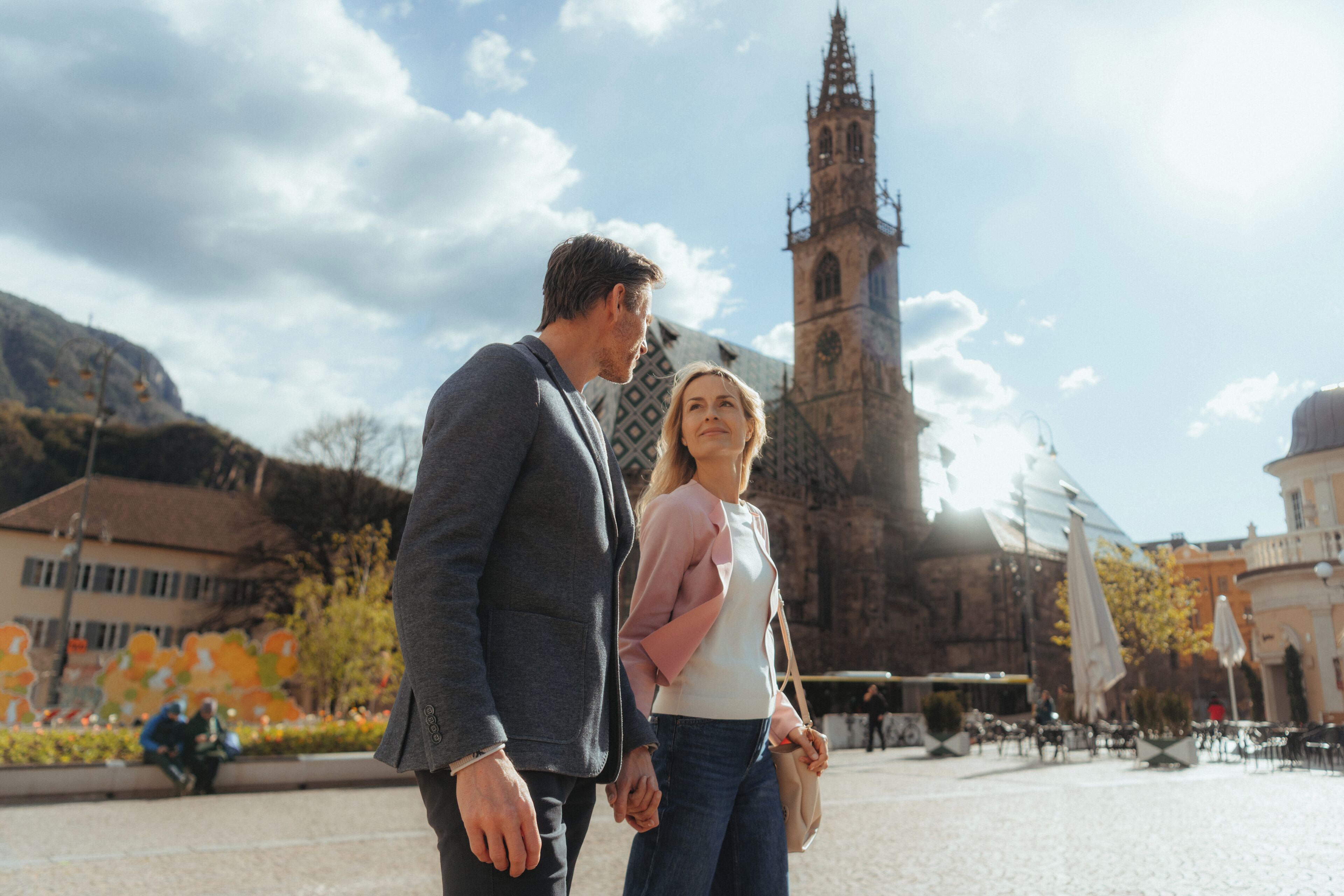 Una pareja tomándose de las manos en una plaza pública con una torre histórica al fondo.