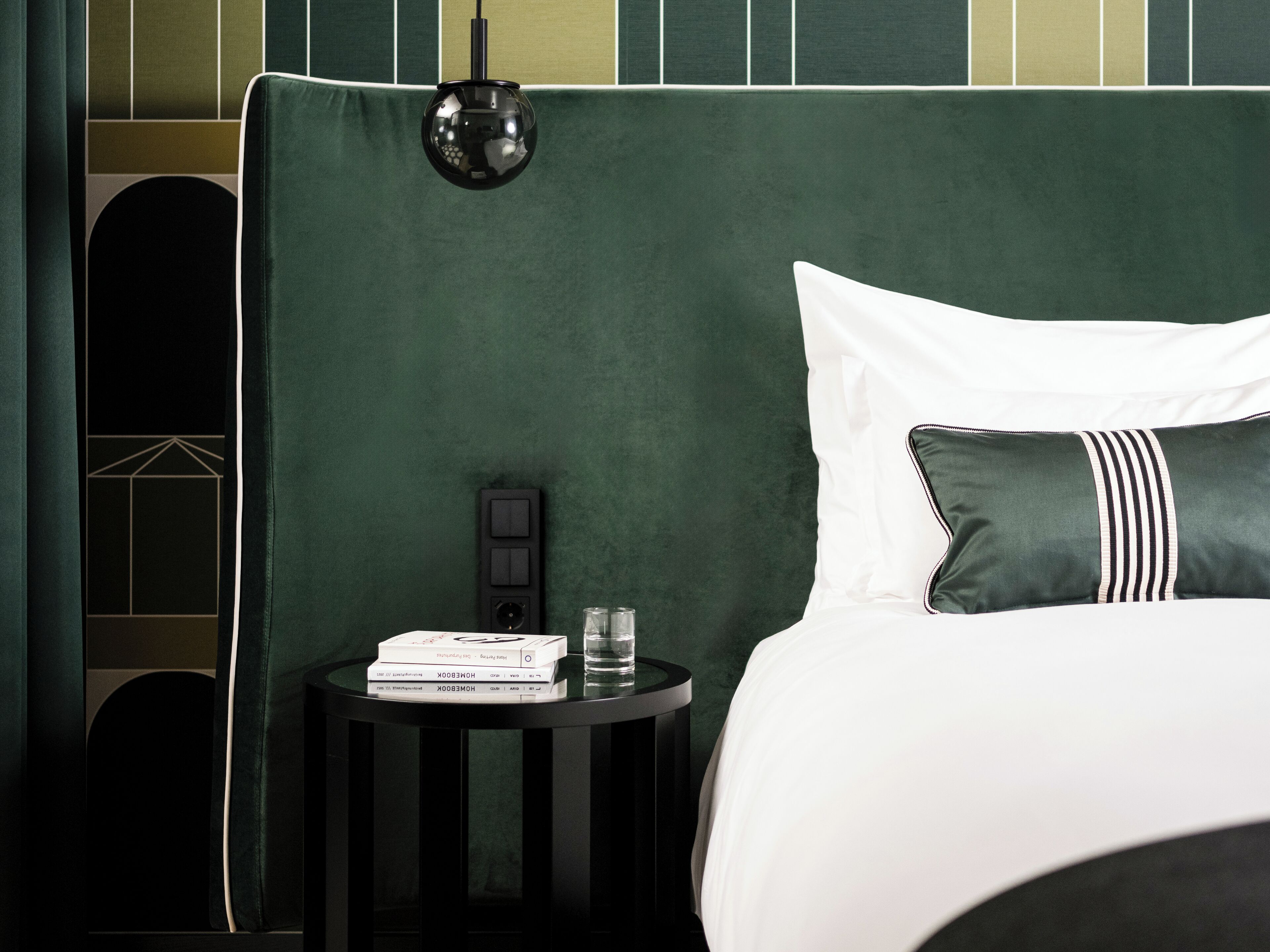A neatly made bed with a green headboard, a bedside table with a lamp, and a stack of books.