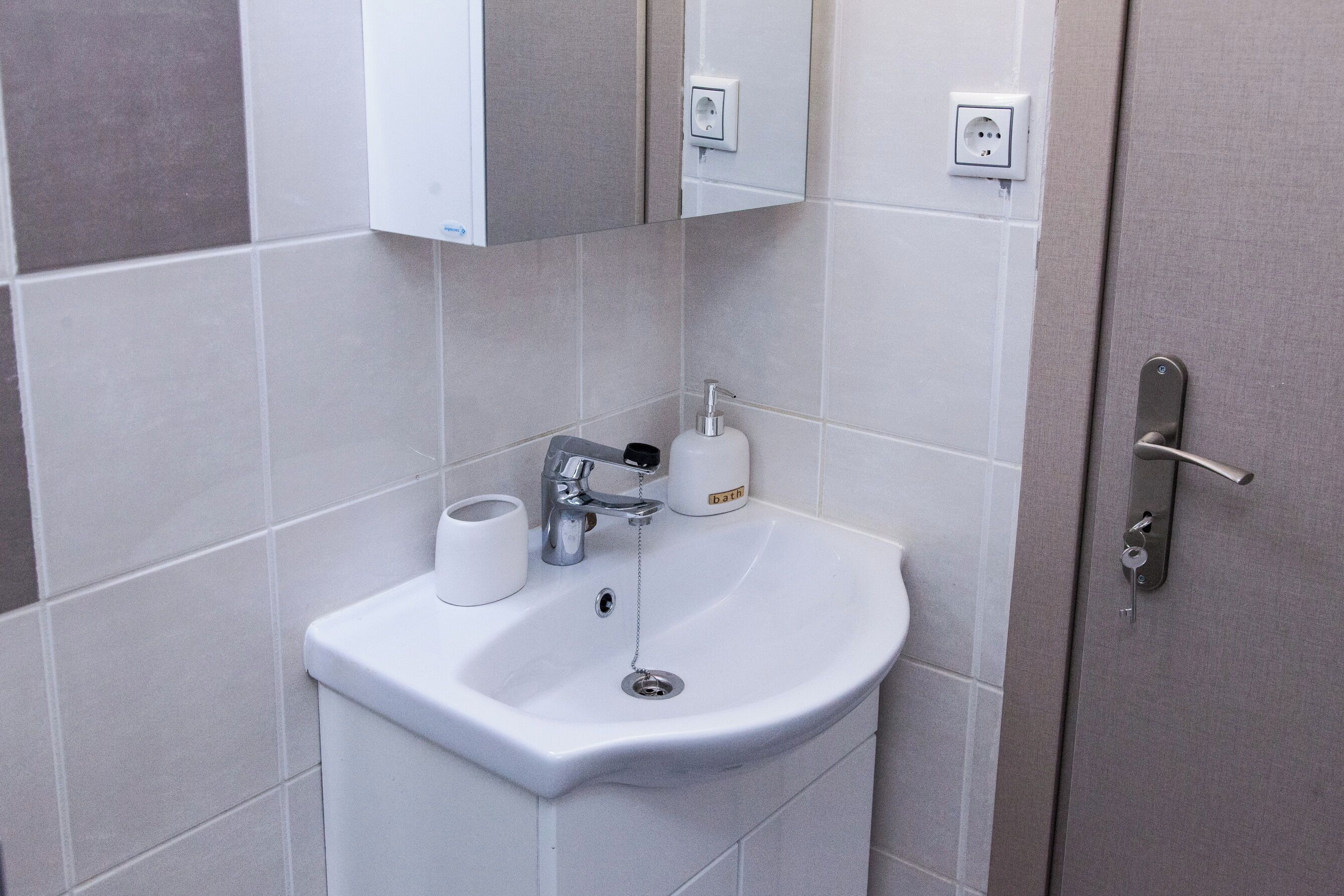 Ein Badezimmer mit einem weißen Waschbecken, einer Toilette und einem Spiegel über dem Waschbecken. An der Wand befinden sich ein Handtuchspender und ein Seifenspender.