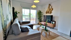 A modern living room with a grey sofa, a coffee table, and a TV on a wooden cabinet. There are plants and a wall art piece.