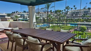 A terrace with a wooden table and wicker chairs, overlooking a pool and a landscaped area with palm trees. - Spacious Apartment with Terrace near Beach Puerto Banus (Marbella)