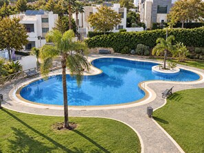 A swimming pool with a curved design, surrounded by a paved walkway, palm trees, and a well-maintained lawn. - Luxury Townhouse Santa Clara Golf (Marbella)