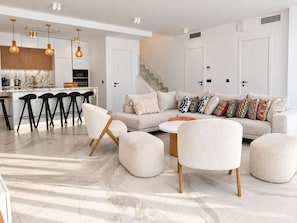 A modern living room with a sofa, ottomans, and a coffee table. There is a kitchen area with bar stools and pendant lighting.
