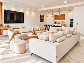 Un salon moderne comprenant un canapé, un pouf et des fauteuils, un téléviseur à écran plat et un coin cuisine avec des tabourets de bar.