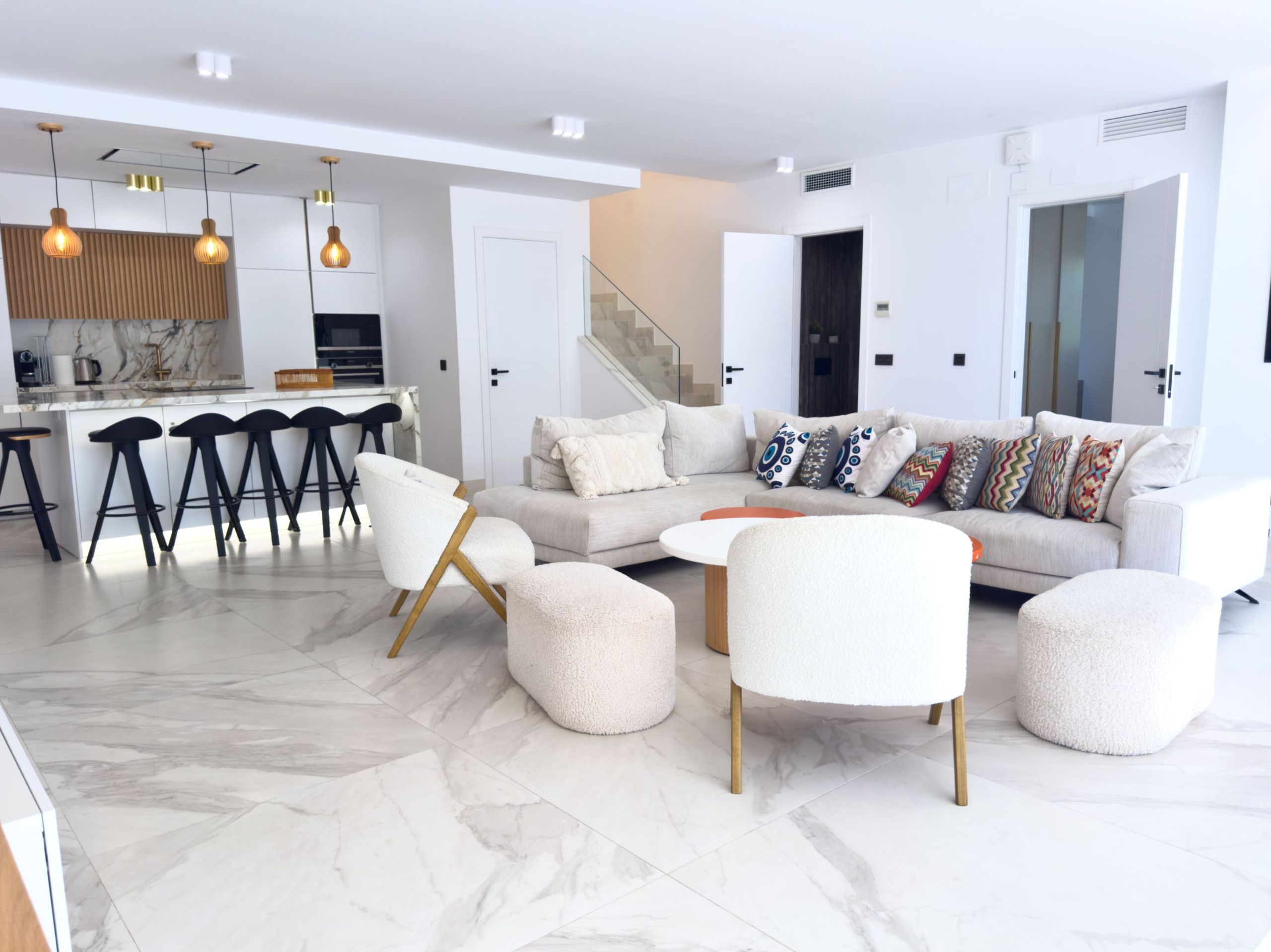 A modern living room with a sofa, ottomans, and a coffee table. There is a kitchen area with bar stools and pendant lighting.