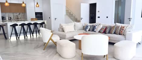Una sala de estar moderna con un sofá, otomanes y una mesa de centro. Hay una zona de cocina con taburetes de bar e iluminación colgante.