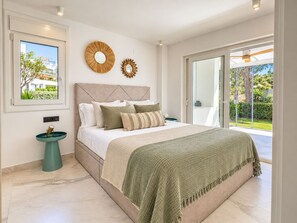 A modern bedroom with a large bed, a view of the outdoors, and a small side table. - Luxury Townhouse Santa Clara Golf (Marbella)