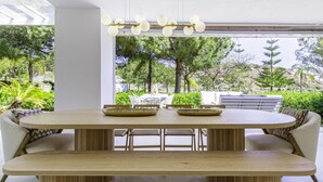 A modern dining area with a wooden table and benches, a ceiling light fixture, and a view of a lush garden. - Luxury Townhouse Santa Clara Golf (Marbella)