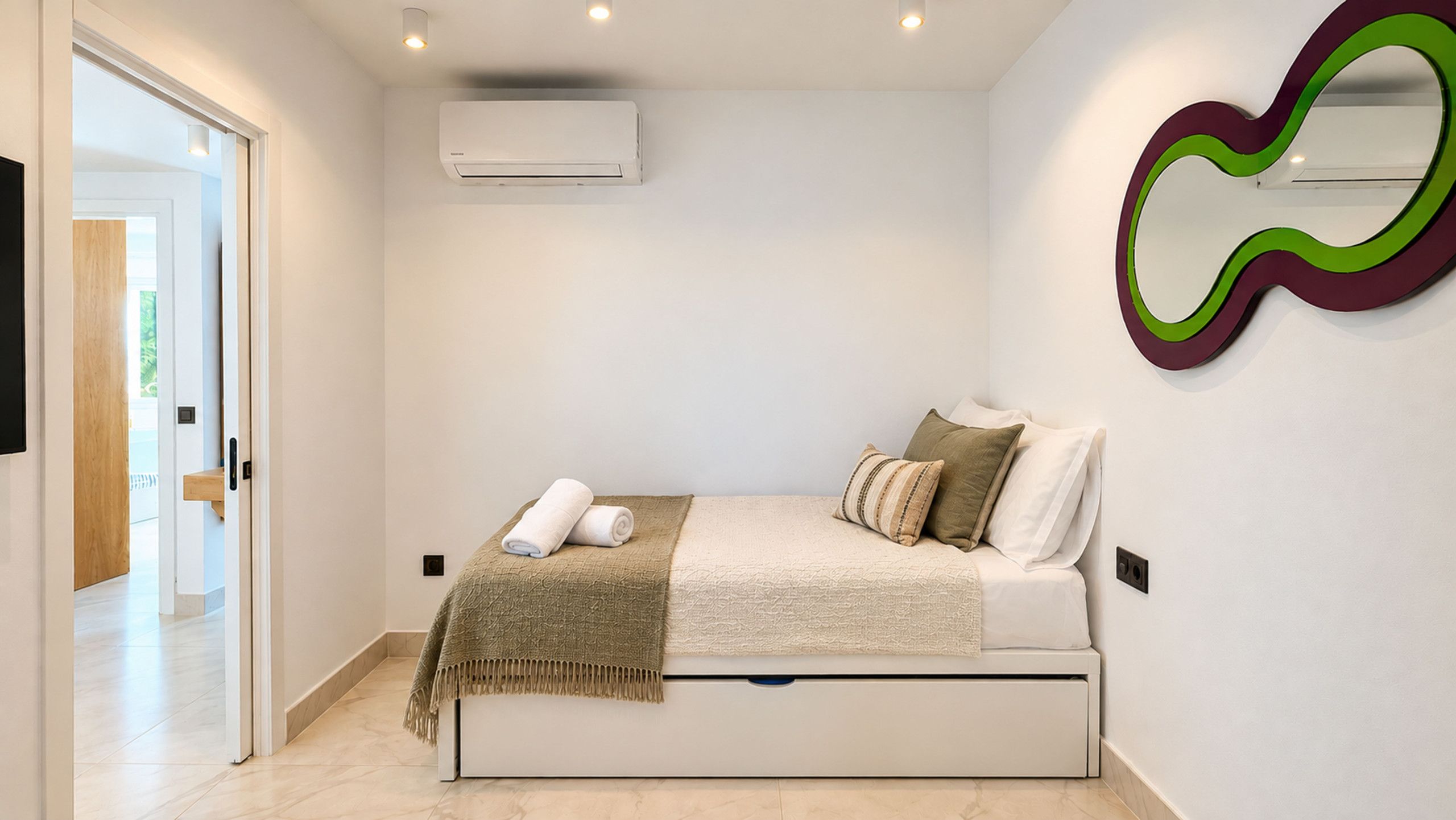 A bedroom with a bed, pillows, a blanket, an air conditioner, and a wall-mounted mirror.