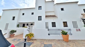 Un bâtiment blanc avec de nombreuses fenêtres et un toit recouvert de tuiles. On y voit des plantes en pot et une rambarde en métal à l’avant.