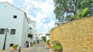 Une ruelle étroite pavée, bordée de bâtiments blancs aux petites fenêtres et d’un mur de pierre d’un côté.