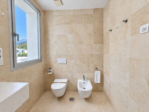 A modern bathroom with a toilet, bidet, and sink, featuring beige tiles and a window with a view of mountains.