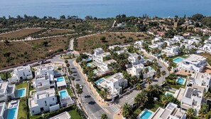An aerial view of a coastal residential area with modern white buildings, swimming pools, and a clear view of the sea.