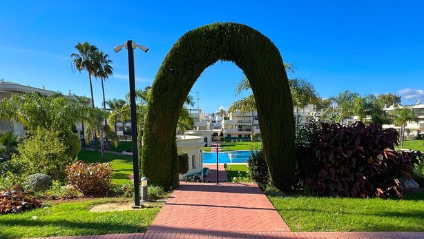 Un sendero conduce a una zona de piscina con un arco de follaje, rodeado de vegetación cuidada y edificios al fondo.
