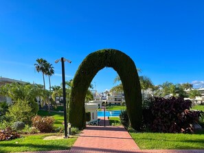 Un chemin mène à un espace piscine agrémenté d’une arche ornée de sculptures végétales, le tout étant entouré d’une végétation luxuriante et bien entretenue, avec des bâtiments en arrière-plan.