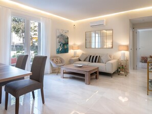 Una sala moderna con un sofá, una mesa de centro y sillas. Hay una mesa de comedor con sillas, una lámpara y un cuadro en la pared.