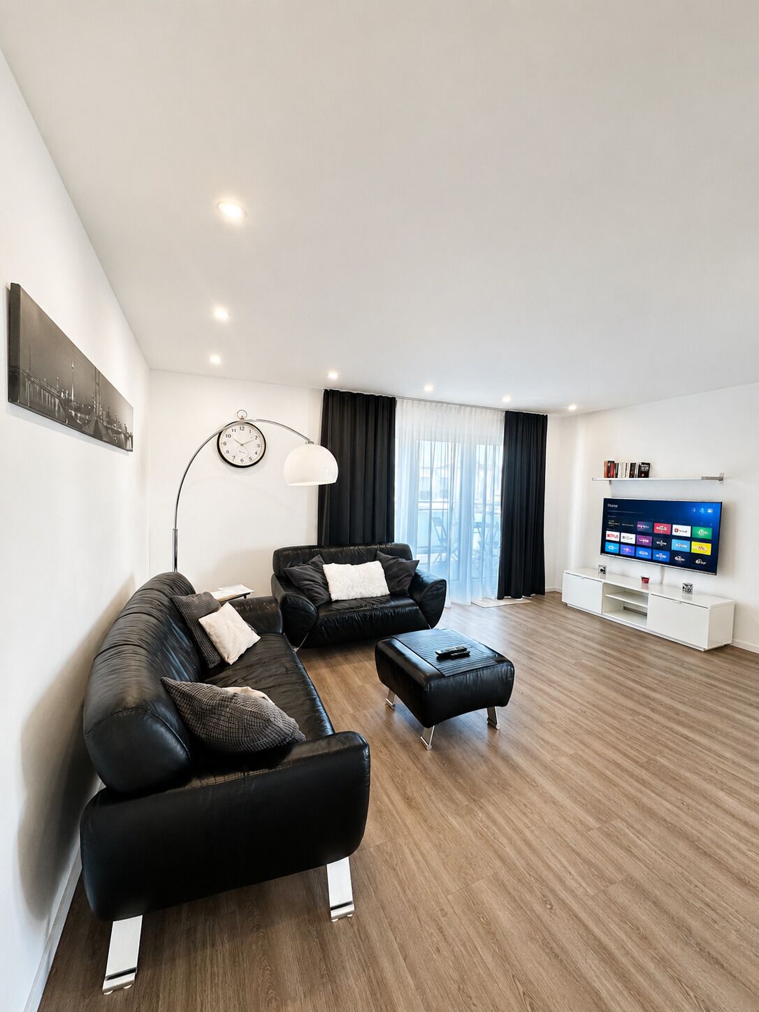 Een moderne woonkamer met een zwarte leren bank, een salontafel, een vloerlamp en een flatscreen televisie op een witte standaard.