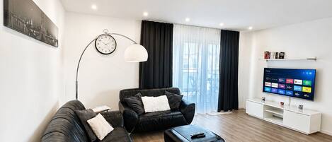 Un salon moderne avec un canapé en cuir noir, une table basse, un lampadaire et un téléviseur à écran plat posé sur un meuble blanc.