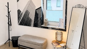 Une pièce au design moderne, agrémentée d’une grande œuvre d’art représentant un paysage urbain encadré, d’un banc, d’une petite table avec une lampe et d’un grand miroir.