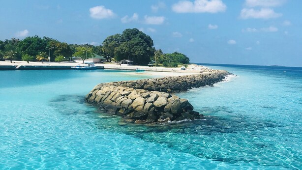 A clear blue sea with a rocky outcrop in the foreground and a sandy beach with trees in the background. - Bon Abri maldives (Magoodhoo)