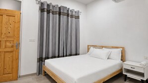 A bedroom with a wooden bed, white bedding, a grey curtain, a white nightstand, and an air conditioning unit. - Bon Abri maldives (Magoodhoo)