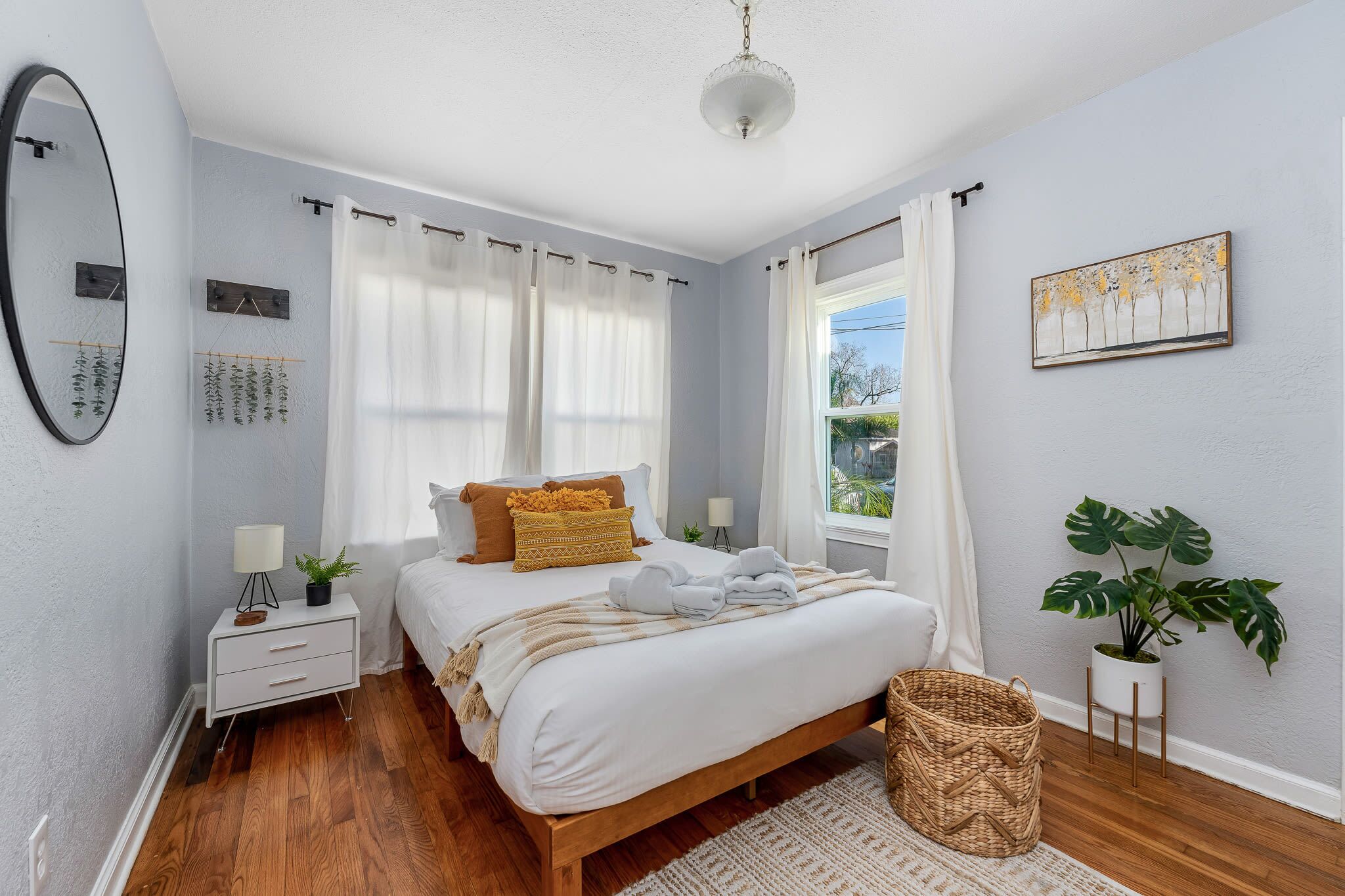Une chambre à coucher avec un lit en bois, une literie blanche, une table de chevet avec une lampe, un miroir fixé au mur et une fenêtre avec des rideaux.