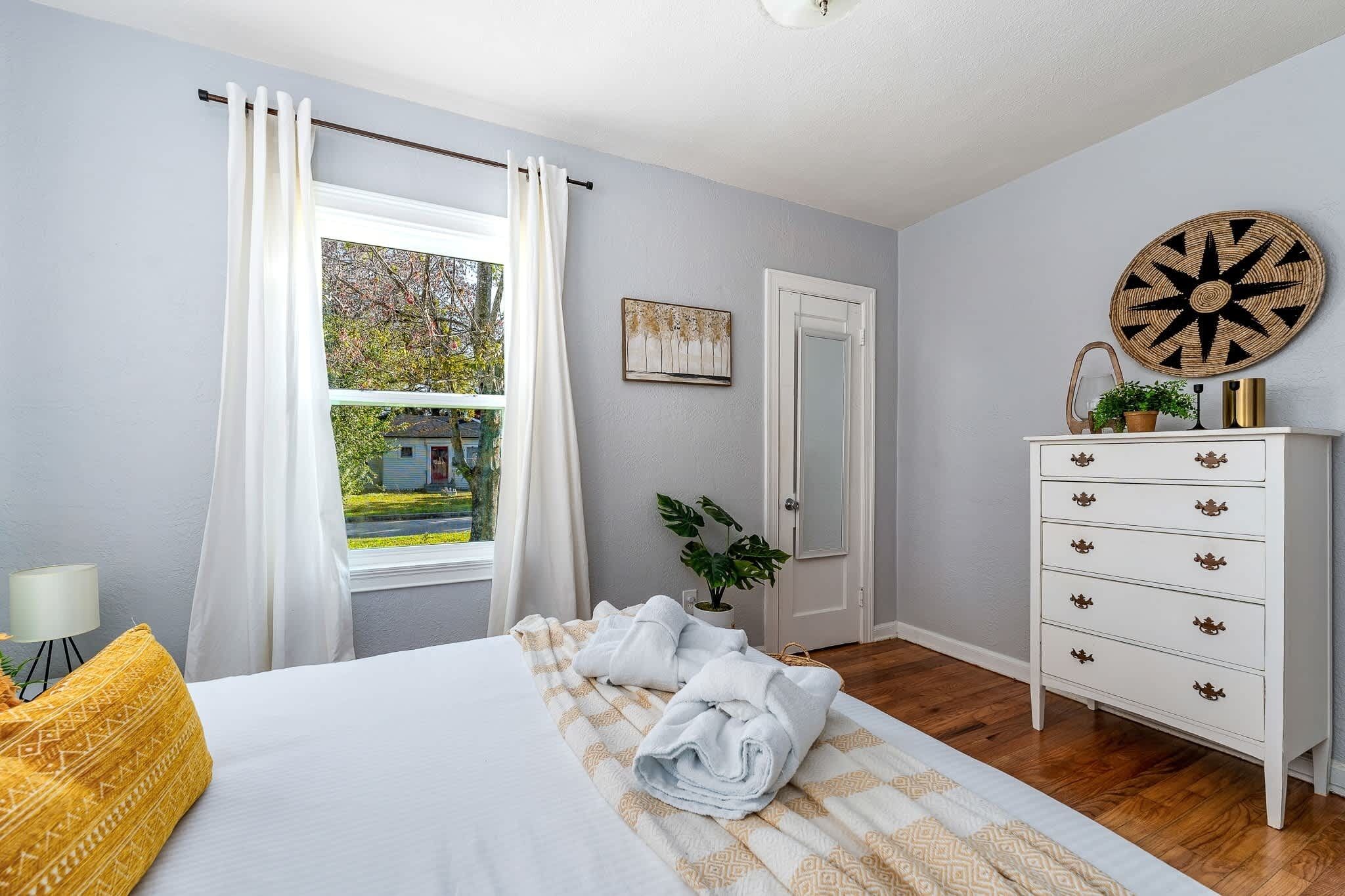 A neatly made bed with white linens, a white dresser with drawers, a round wall decoration, and a window with curtains.