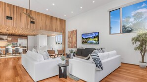 A modern living room with a large white sectional sofa, a flat-screen TV mounted on the wall, and a dining area with a wooden ceiling. - Cedar Loft Villa (Venice)