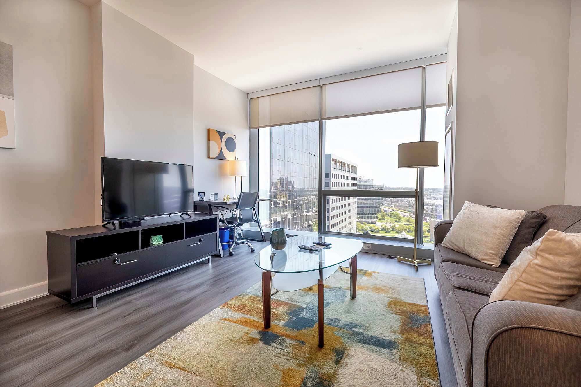 A modern living room with a flat-screen TV, a glass coffee table, a sofa, and a large window offering a city view.