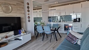 A modern kitchen with white cabinets, a dining area with blue chairs, and a flat-screen TV on a white console. - SUNSET VIEW HILL (Costa Adeje)