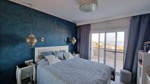 A bedroom with a large bed, a nightstand, a dresser, and a view of the city through a balcony. - SUNSET VIEW HILL (Costa Adeje)