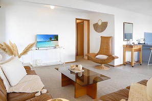 A modern living room with a sofa, a glass coffee table, a chair, a television, and a kitchen area with a coffee machine.