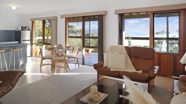 A modern living room with a brown leather sofa, a dining area with a table and chairs, and large windows offering a view of the outdoors.