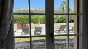 Une vue à travers une fenêtre donnant sur un mur de pierre et deux fauteuils de jardin placés près d’une piscine.