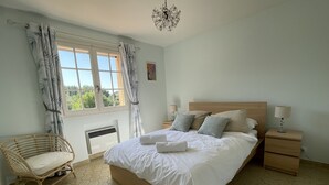 Une chambre à coucher avec un lit en bois, du linge de lit blanc, une chaise en osier, une table de chevet avec une lampe et une fenêtre avec des rideaux.