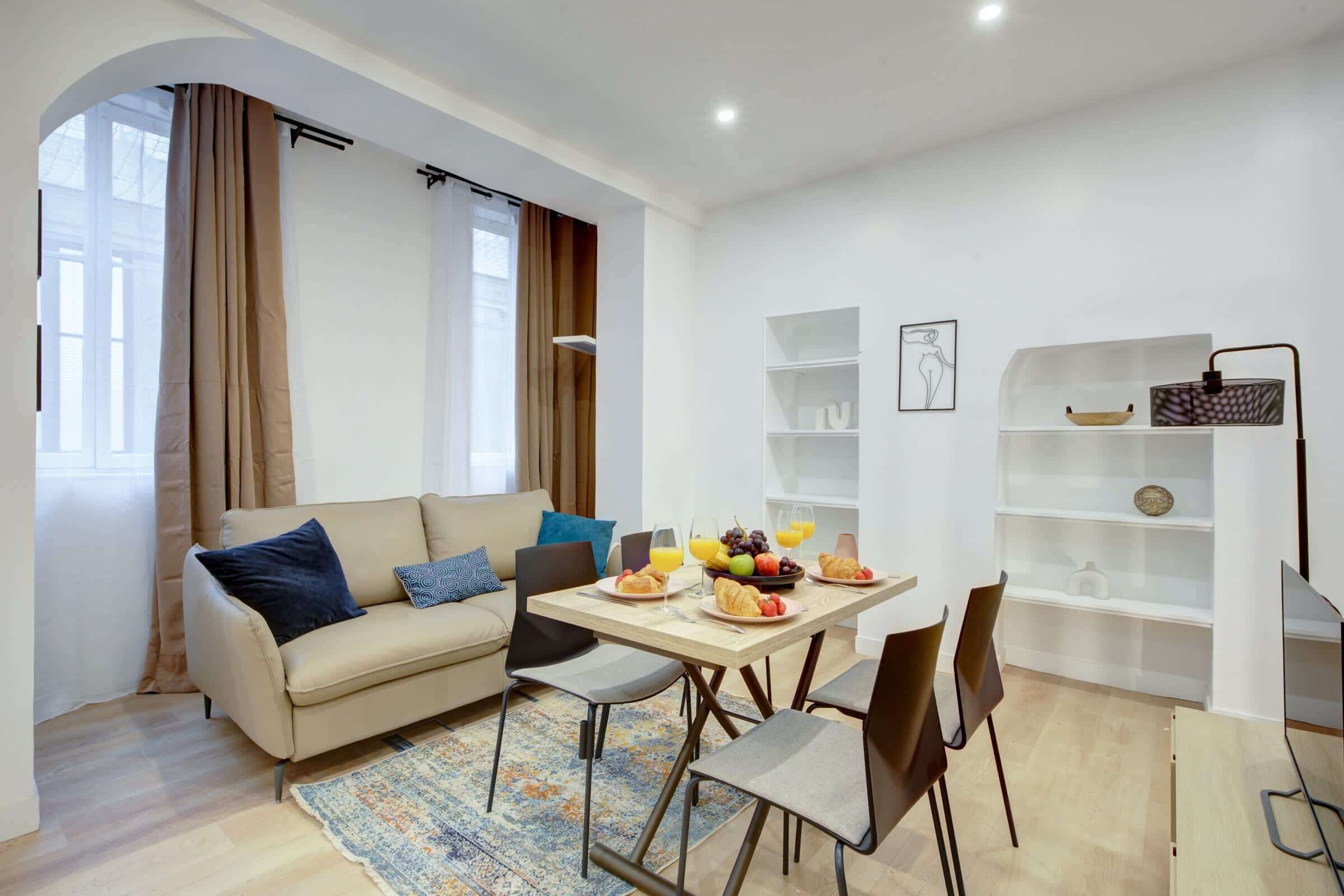 A modern living room with a sofa, dining table, and chairs. There is a breakfast setup with fruits and a bowl of cereal.