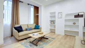 A modern living room with a beige sofa, a coffee table, and a patterned rug. There are white shelves with decorative items and a framed picture on the wall. - Charming 1BR in Montorgueil - Les Halles (Paris)