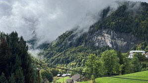 Paisaje montañoso con una valla de madera en primer plano, un campo cubierto de hierba y algunos edificios a lo lejos.