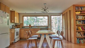 A modern kitchen with wooden cabinets, a dining table with chairs, a refrigerator, and a bookshelf filled with books. - Quiet, cozy home in the woods CS0038 (Wellfleet)