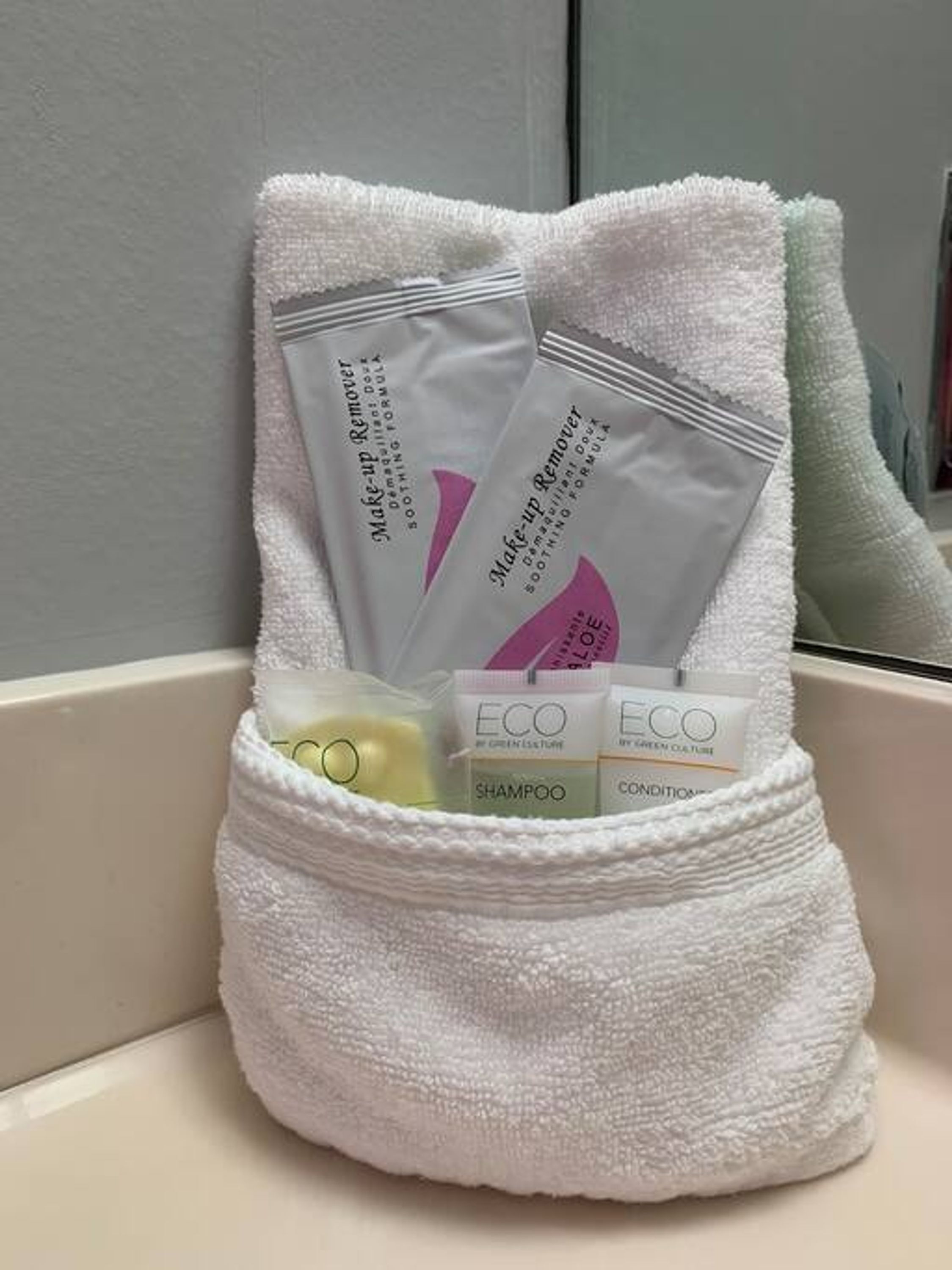 A white towel with a pocket containing a make-up remover, a shampoo, and a conditioner.