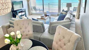 A spacious living room with a large sofa set, a coffee table, and a TV. There is a view of the ocean through large windows. - Ocean Front Condo in New Smyrna Beach FL (New Smyrna Beach)