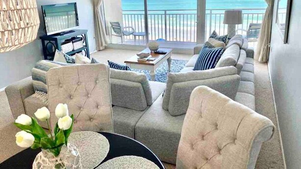 A spacious living room with a large sofa set, a coffee table, and a TV. There is a view of the ocean through large windows. - Ocean Front Condo in New Smyrna Beach FL (New Smyrna Beach)