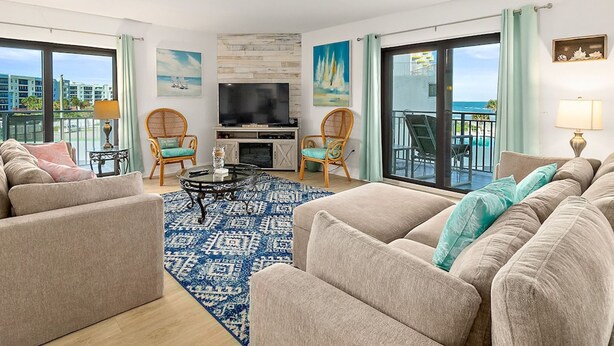 A spacious living room with a large flat-screen TV, comfortable seating, a patterned rug, and large windows offering a view of the ocean and buildings. - Oceanview 3 Bedroom Condo New Smyrna Beach FL (New Smyrna Beach)
