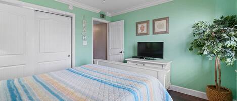 A bedroom with a bed, a ceiling fan, a television, a plant, and a door.
