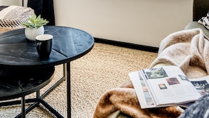 Una habitación moderna con un ventanal grande, una mesa redonda negra con una planta y una taza, un plaid acogedor y una revista sobre la cama.