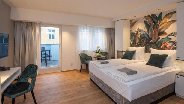 Une chambre d’hôtel avec un lit, un bureau, une chaise, une petite table avec un vase et une grande fenêtre avec des rideaux.