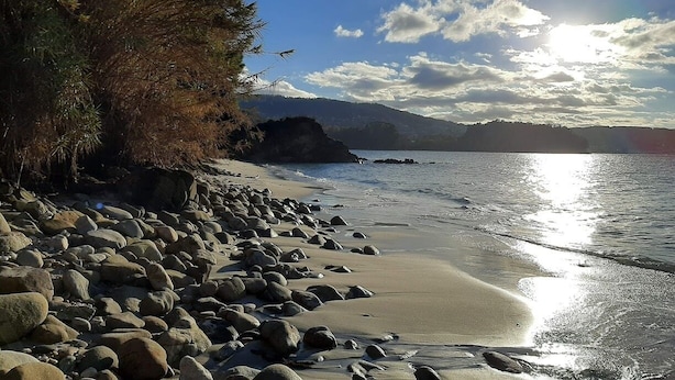 Ein felsiger Strand mit klarem Himmel und Sonnenlicht, das sich im Wasser spiegelt.