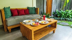 A wooden sofa with green and red cushions, a wooden coffee table with plates of food, and a glass of juice. - Sawitri Anandhita Luxury Villas (Ubud)
